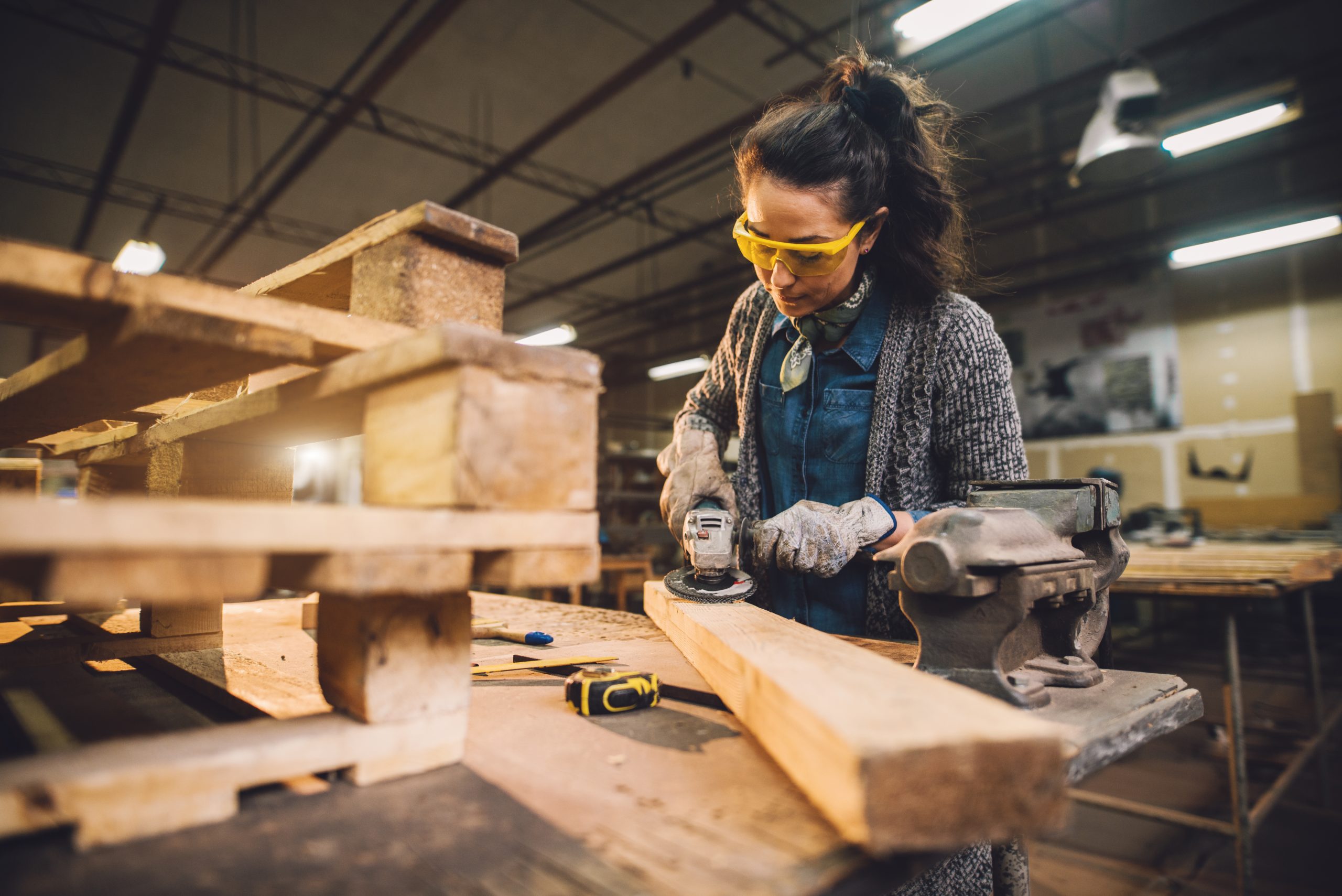 Bild einer Frau mit Schutzbrille in einer Werkstatt welche Schleifarbeiten an einem Holzbalken durchführt. Beispielbild der Holzindustrie.